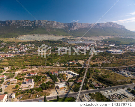 Railway line runs through rural valley beneath rugged mountains. Elevated track connects scattered houses and industrial sites, with broad quarry cut into forested slopes beyond, winding along Railway line runs through rural valley beneath rugged mountains. Elevated track connects scattered houses and industrial sites, with broad quarry cut into forested slopes beyond, winding along 143379771