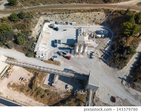 Aerial view of cement plant with trucks, silos, and conveyor system across dusty industrial yard. Concrete production facility sits among scrubby hills, with aggregate piles and access roads. Aerial view of cement plant with trucks, silos, and conveyor system across dusty industrial yard. Concrete production facility sits among scrubby hills, with aggregate piles and access roads. 143379780