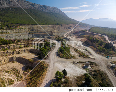 Mountain quarry occupies broad hillside, with stepped excavation benches and pale dirt roads across its floor. Forested slopes frame open mine, with excavator and tanks nearby, beside terraced ground. 143379783