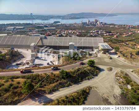 Cement factory occupies foreground beside broad coastal bay and urban shoreline. Aerial perspective reveals processing buildings, conveyors, storage silos, and haul trucks on dusty access roads. Cement factory occupies foreground beside broad coastal bay and urban shoreline. Aerial perspective reveals processing buildings, conveyors, storage silos, and haul trucks on dusty access roads. 143379784