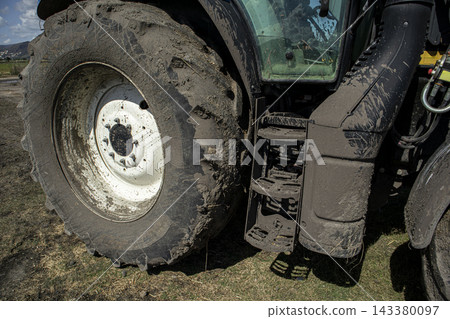 Heavy tractor wheel covered in mud parked near grassy field Heavy tractor wheel covered in mud parked near grassy field 143380097
