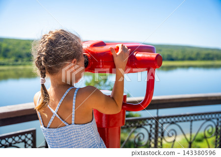 Young girl looking through viewing telescope at river landscape. Family travel encourages children to explore nature and discover lesser known destinations 143380596