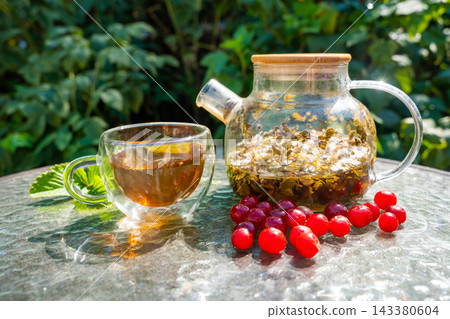 Glass cup and teapot with herbal tea served with fresh cherries and garden leaves. Homemade infusion represents preparing drinks from garden plants and berries Glass cup and teapot with herbal tea served with fresh cherries and garden leaves. Homemade infusion represents preparing drinks from garden plants and berries 143380604