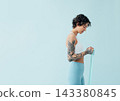 Side view of a slim woman in sportswear practicing with a resistance band over a blue backdrop 143380845