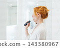 Middle aged woman cleaning her teeth with electronic toothbrush in her white modern bathroom. Using Sonic toothbrush for routine oral care. Modern home health care technology concept. 143380967