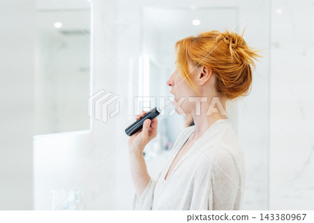 Middle aged woman cleaning her teeth with electronic toothbrush in her white modern bathroom. Using Sonic toothbrush for routine oral care. Modern home health care technology concept. Middle aged woman cleaning her teeth with electronic toothbrush in her white modern bathroom. Using Sonic toothbrush for routine oral care. Modern home health care technology concept. 143380967