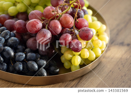 Assortment of fresh red green and black grapes displayed in a round brass plate on a wooden surface Assortment of fresh red green and black grapes displayed in a round brass plate on a wooden surface 143381569