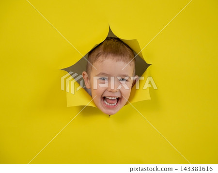 A mischievous boy peeks out through a hole in a yellow cardboard background. A mischievous boy peeks out through a hole in a yellow cardboard background. 143381616
