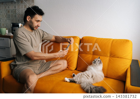 Happy young man sitting on bright orange sofa photographing fluffy cat with smartphone. Relaxed domestic lifestyle scene showing human animal bond and social media content creation at home. Happy young man sitting on bright orange sofa photographing fluffy cat with smartphone. Relaxed domestic lifestyle scene showing human animal bond and social media content creation at home. 143381732