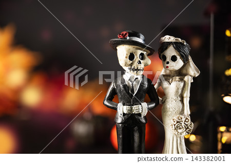 Skeleton bride and groom figurines beside glowing haunted house, pumpkins and smoke. Gothic Halloween wedding decorations on dark background with ample copy space. Halloween holiday card. Skeleton bride and groom figurines beside glowing haunted house, pumpkins and smoke. Gothic Halloween wedding decorations on dark background with ample copy space. Halloween holiday card. 143382001