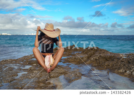 Woman in straw hat sitting on coastal rock holding pink conch shell Woman in straw hat sitting on coastal rock holding pink conch shell 143382426
