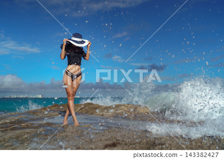 Woman in wide brim hat on rocky shore with splashing ocean wave 143382427