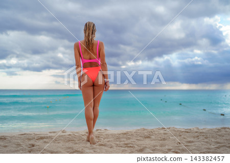 Woman in bright swimsuit standing on beach facing turquoise sea 143382457