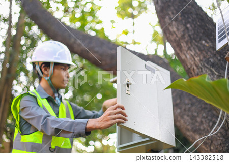 An electrician wearing a white hard hat and a high-visibility vest is installing or inspecting an electrical panel or switch control box out door. An electrician wearing a white hard hat and a high-visibility vest is installing or inspecting an electrical panel or switch control box out door. 143382518