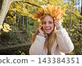 Young woman wearing a wreath of colorful maple leaves relaxes in an autumn park. Seasonal lifestyle portrait celebrating fall colors, outdoor wellness and the beauty of nature. 143384351
