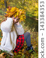 Young woman wearing a wreath of colorful maple leaves relaxes in an autumn park. Seasonal lifestyle portrait celebrating fall colors, outdoor wellness and the beauty of nature. 143384353