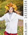 Young woman wearing a wreath of colorful maple leaves relaxes in an autumn park. Seasonal lifestyle portrait celebrating fall colors, outdoor wellness and the beauty of nature. 143384354