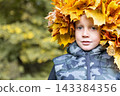 Portrait of a young boy wearing a handmade maple leaf wreath in an autumn park. Fall lifestyle, colorful foliage, outdoor childhood, cozy weekend walk and seasonal nature. 143384356
