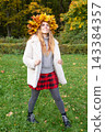 Young woman wearing a wreath of colorful maple leaves relaxes in an autumn park. Seasonal lifestyle portrait celebrating fall colors, outdoor wellness and the beauty of nature. 143384357