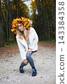 Happy young woman wearing a maple leaf wreath enjoys an autumn park with open arms. Fall lifestyle, colorful foliage, outdoor leisure, wellness and seasonal nature during a weekend walk. 143384358