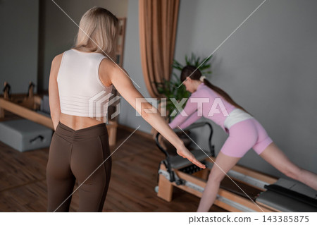 Young caucasian women exercising Scooter on pilates reformers for the hips and glutes in a bright studio. Her workout focuses on strength and flexibility Young caucasian women exercising Scooter on pilates reformers for the hips and glutes in a bright studio. Her workout focuses on strength and flexibility 143385875