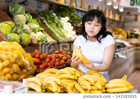 Attentive young woman purchaser choosing bananas in grocery store Attentive young woman purchaser choosing bananas in grocery store 143385935