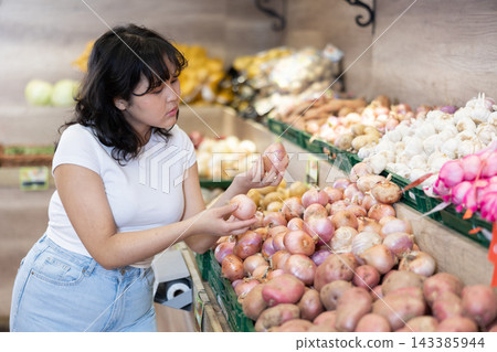Attentive young woman purchaser choosing onions in grocery store Attentive young woman purchaser choosing onions in grocery store 143385944