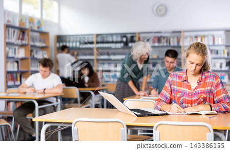 Teenager female student with laptop sitting at desk in library at lesson in high school Teenager female student with laptop sitting at desk in library at lesson in high school 143386155