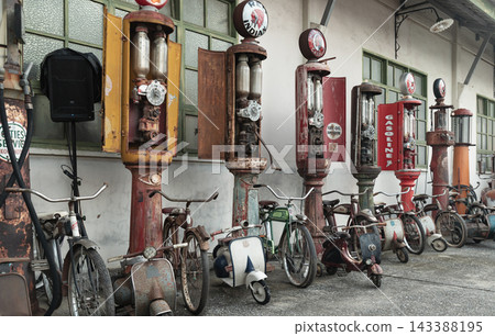 A line of weathered, vintage gasoline pumps with visible glass measuring cylinders stands outdoors alongside a collection of classic bicycles and motor scooters, evoking a nostalgic Americana A line of weathered, vintage gasoline pumps with visible glass measuring cylinders stands outdoors alongside a collection of classic bicycles and motor scooters, evoking a nostalgic Americana 143388195