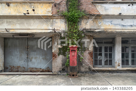 A vintage red post box stands centrally against a heavily weathered brick building facade, framed by creeping green vines scaling the distressed plaster wall. Flanked by a rusted metal garage door on 143388205