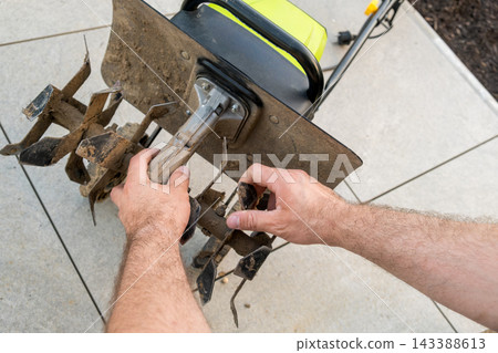 A person is adjusting the tines of a garden tiller on a patio surface. Hands are seen modifying the attachment of a tiller used for cultivating soil in an outdoor setting. A person is adjusting the tines of a garden tiller on a patio surface. Hands are seen modifying the attachment of a tiller used for cultivating soil in an outdoor setting. 143388613