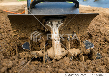 The sharp blades of a tiller are actively working through dark, moist soil, ensuring optimal conditions for vegetable planting. The sharp blades of a tiller are actively working through dark, moist soil, ensuring optimal conditions for vegetable planting. 143388616