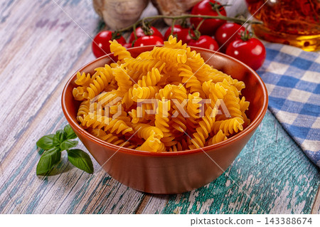 Uncooked fusilli pasta in terracotta bowl with cherry tomatoes basil and garlic on rustic wooden surface food preparation Uncooked fusilli pasta in terracotta bowl with cherry tomatoes basil and garlic on rustic wooden surface food preparation 143388674