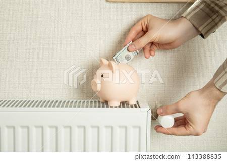 Heating season. Hand places cash in piggy bank on radiator. Budgeting for heating season concept Heating season. Hand places cash in piggy bank on radiator. Budgeting for heating season concept 143388835