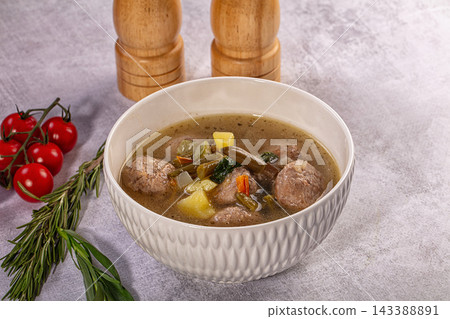 White ceramic bowl filled with meatball soup containing potatoes green beans carrots and herbs on textured grey surface with cherry tomatoes and rosemary sprig nearby food dish White ceramic bowl filled with meatball soup containing potatoes green beans carrots and herbs on textured grey surface with cherry tomatoes and rosemary sprig nearby food dish 143388891