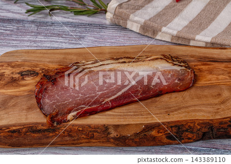 Rustic wooden board holding cured prosciutto slice alongside rosemary sprig and striped kitchen towel Rustic wooden board holding cured prosciutto slice alongside rosemary sprig and striped kitchen towel 143389110