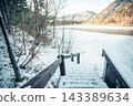 Wooden staircase descending through a snowy forest toward a frozen mountain lake surrounded by rocky peaks. Winter landscape with fresh snow, bare trees, tranquil atmosphere and scenic wilderness 143389634
