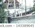 closeup of rustic wooden house with green roof and stone fence in snowy winter forest, birch trees and wooden railing, countryside lifestyle, tourism and seasonal recreation 143389645