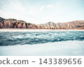 winter river flows through a snowy landscape. Mountains rise in the background, surrounded by bare trees. The scene evokes a sense of peace and mindfulness. 143389656