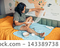 Young woman sitting on a bed while changing a bandage on her injured leg at home. Medical supplies, gauze, scissors, and home care items illustrate wound treatment, first aid, rehabilitation, and 143389738