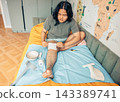 plus size young woman applying a medical gauze dressing during first aid practice at home while sitting on a bed with healthcare supplies and a crutch nearby. concept of healthcare, rehabilitation 143389741