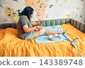 woman sitting on a bed and applying gauze bandage to her injured leg at home. visible scars and bruising on the knee and thigh. home first aid, wound care, injury recovery, rehabilitation, self care 143389748