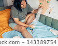 plus size young woman sitting on a bed and applying a gauze bandage to her injured leg at home. visible scars and bruising on the knee and thigh. home first aid, wound care, injury recovery 143389754