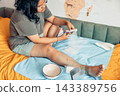 plus size young woman sitting on a bed and cutting gauze for dressing an injured leg at home. visible scars and healing wounds on her knee and lower leg. home first aid, wound care, injury recovery 143389756