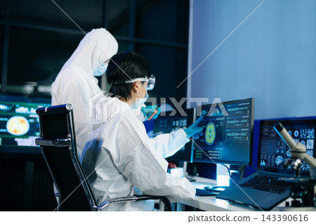 Professional scientists in protective suits collaborating on research, examining microscope samples, and recording data Professional scientists in protective suits collaborating on research, examining microscope samples, and recording data 143390616