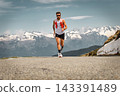 Athletic bearded man running on a mountain road wearing trail running gear against a scenic alpine landscape background Athletic bearded man running on a mountain road wearing trail running gear against a scenic alpine landscape background 143391489