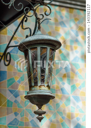 Ornate metal lantern hanging from a black wrought iron bracket against a vibrant mosaic wall 143392117