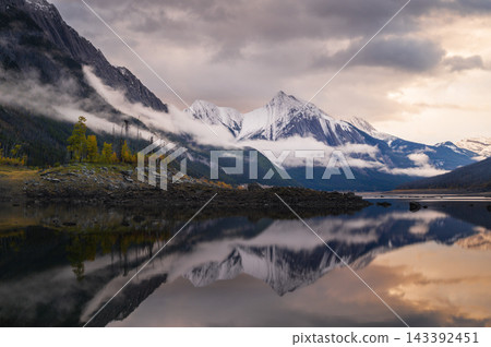 Tranquil mountain landscape with snow reflection in calm lake water and scenic cloud over peaceful wild nature of majestic canada Tranquil mountain landscape with snow reflection in calm lake water and scenic cloud over peaceful wild nature of majestic canada 143392451