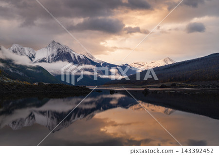 Serene canada mountain wilderness offering peaceful reflection on calm lake water beneath snow peak landscape during dramatic golden sunset Serene canada mountain wilderness offering peaceful reflection on calm lake water beneath snow peak landscape during dramatic golden sunset 143392452