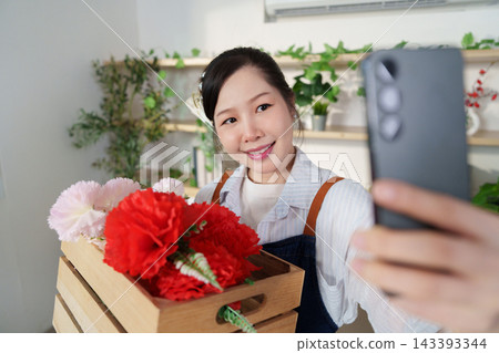 Asian florist taking selfie with smartphone holding crate of carnations Asian florist taking selfie with smartphone holding crate of carnations 143393344
